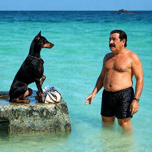 Laurent Gérard torse nu dans l'eau turquoise aux côtés d'un doberman, ambiance Magnum P.I.